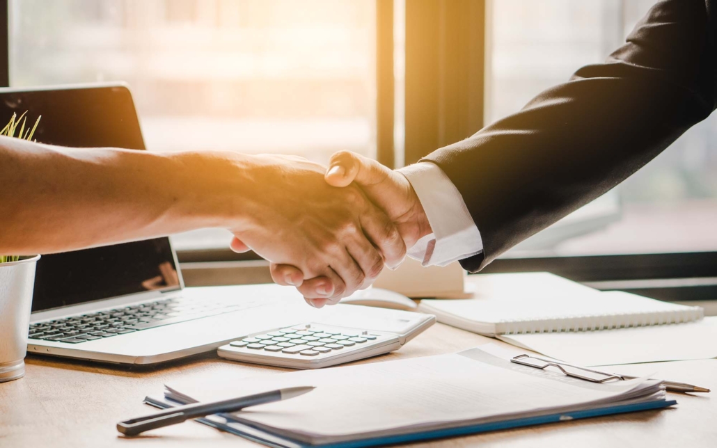 Two individuals are shaking hands across a desk, symbolizing a successful agreement or partnership. The desk features a laptop, a calculator, and documents, emphasizing a business setting. The warm sunlight filtering through the window adds a positive and welcoming ambiance to the professional interaction.