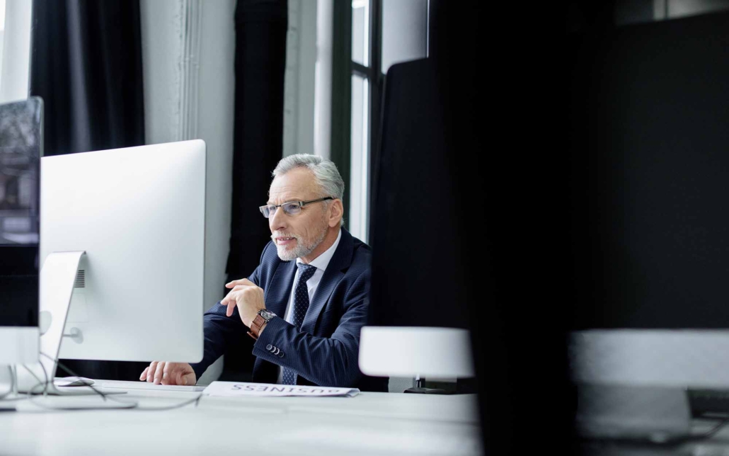 A senior business professional, dressed in a suit, sits at a desk working on a large computer monitor. His focused expression and attentive posture indicate deep engagement with the task at hand, reflecting a blend of experience and dedication in a modern office setting.