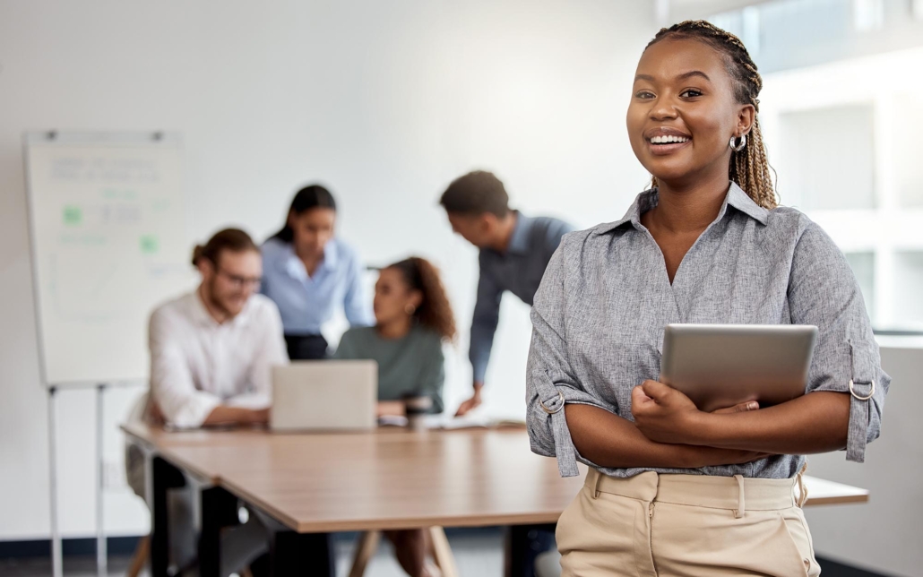A business person holds a tablet and smiles, standing in front while a group of colleagues engages in a meeting in the background. Her positive demeanor and the tablet in hand suggest a role in managing or facilitating the meeting, reflecting a blend of approachability and professionalism.