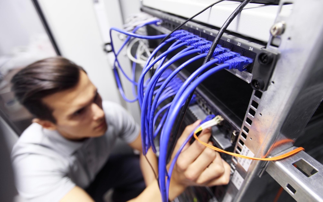 An IT technician works on a network rack, carefully managing and connecting blue ethernet cables. The technician's focused attention and precise handling of the cables emphasize the importance of proper network setup and maintenance to ensure reliable connectivity.