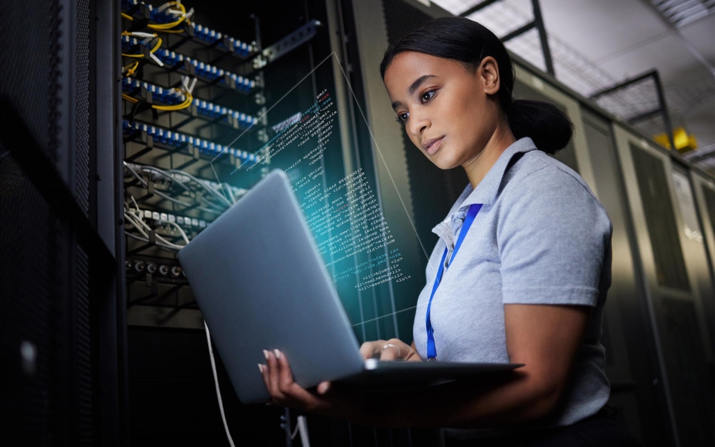 An IT professional works in a server room, focusing intently on her laptop. The surrounding racks of servers and the complex code displayed on her screen indicate a critical task in managing and maintaining the data center's infrastructure. Her concentrated expression underscores the importance of her work in ensuring system stability and security.