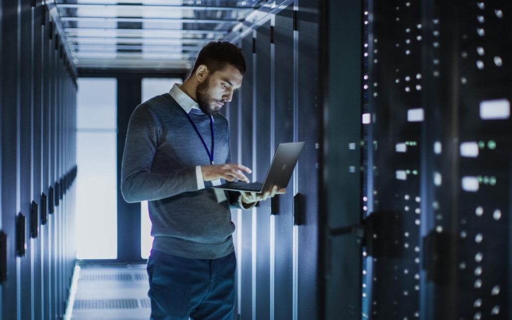 An IT professional works in a dimly lit server room, intently focused on a laptop. Surrounded by rows of servers, the professional's dedicated attention to the screen highlights the critical task of managing and maintaining the data center's infrastructure.