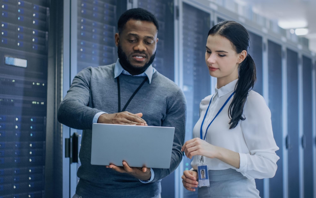 Two IT professionals discuss data and operations while looking at a laptop in a server room. Surrounded by racks of servers, their focused conversation highlights the collaborative effort required to manage and optimize the data center’s performance and security.
