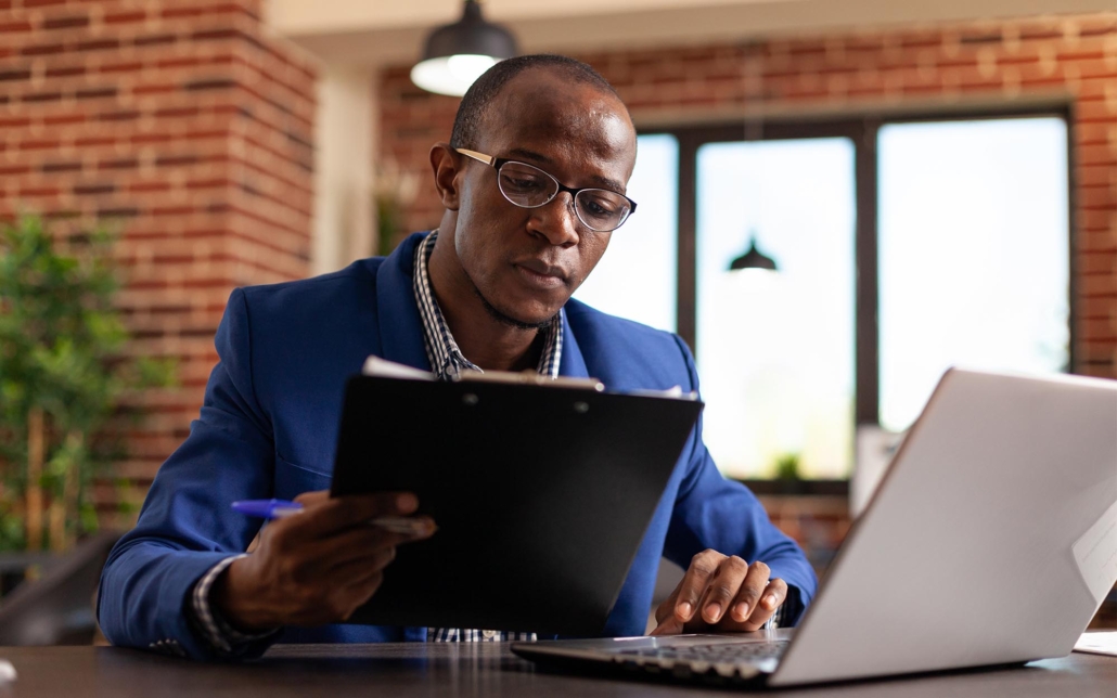 A professional reviews documents on a clipboard while working at a desk with a laptop. His focused expression and careful attention to detail indicate a thorough analysis of the paperwork, reflecting a diligent approach to his tasks.