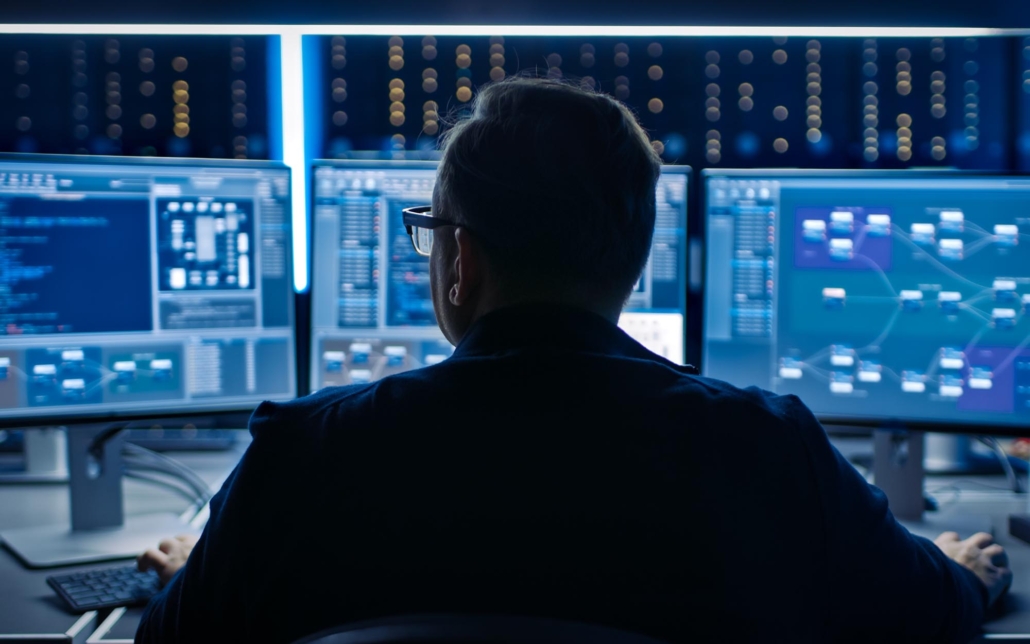 A cybersecurity professional works at a desk with multiple monitors displaying complex data and network diagrams. The dimly lit room and the intense focus on the screens suggest a high-tech environment where monitoring and analyzing network security are paramount.