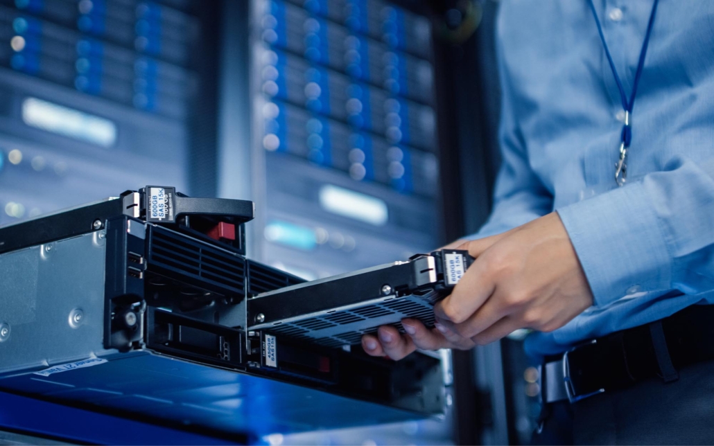 A technician works in a server room, carefully handling a hard drive while performing maintenance on a server. The environment is filled with rows of server racks, indicating a critical task aimed at ensuring the smooth operation and reliability of the data center.
