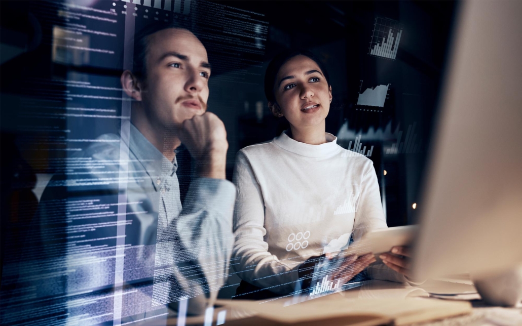Two professionals analyze data on a computer screen, with various graphs and code displayed in front of them. One person thoughtfully rests their chin on their hand while the other explains something, reflecting a collaborative and analytical approach to data interpretation and problem-solving.