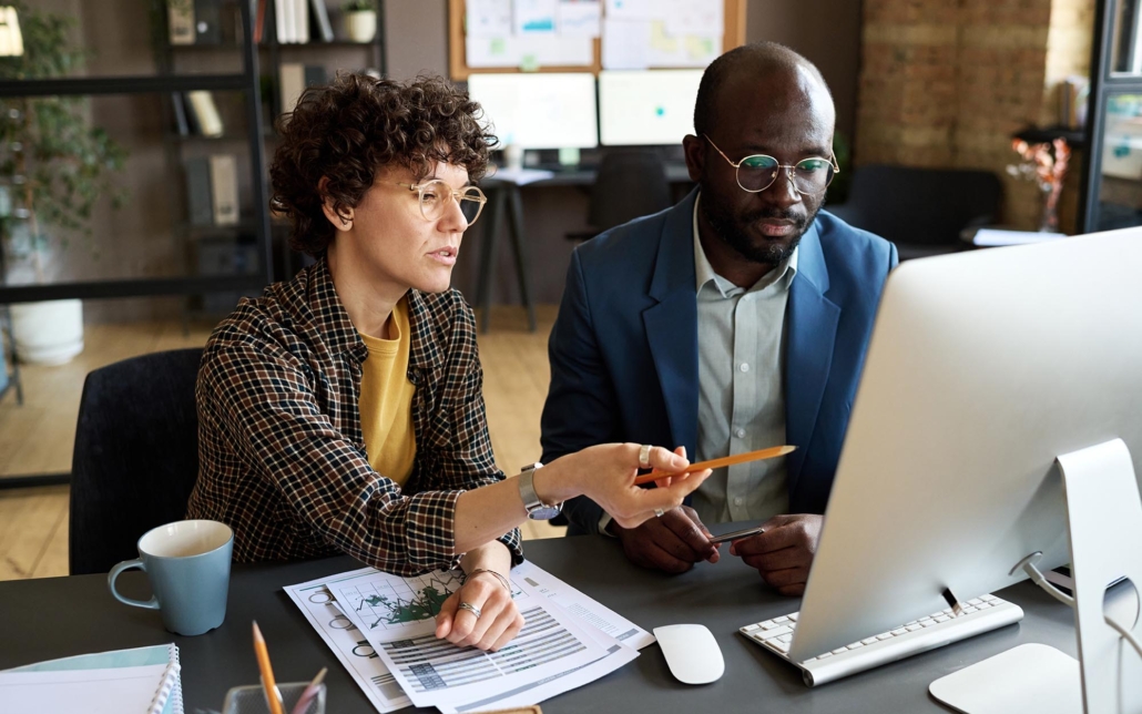 Two colleagues collaborate at a desk, intently looking at a computer screen. One person points to the screen with a pencil, while the other listens attentively. Papers with charts and graphs are spread out on the desk, indicating a focused discussion on data analysis or strategy planning.