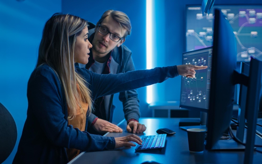 Two professionals work together in a dimly lit control room, analyzing data on multiple computer screens. One person points to the screen while explaining something, and the other listens intently. The screens display complex data and network diagrams, indicating a detailed discussion on network management or cybersecurity.