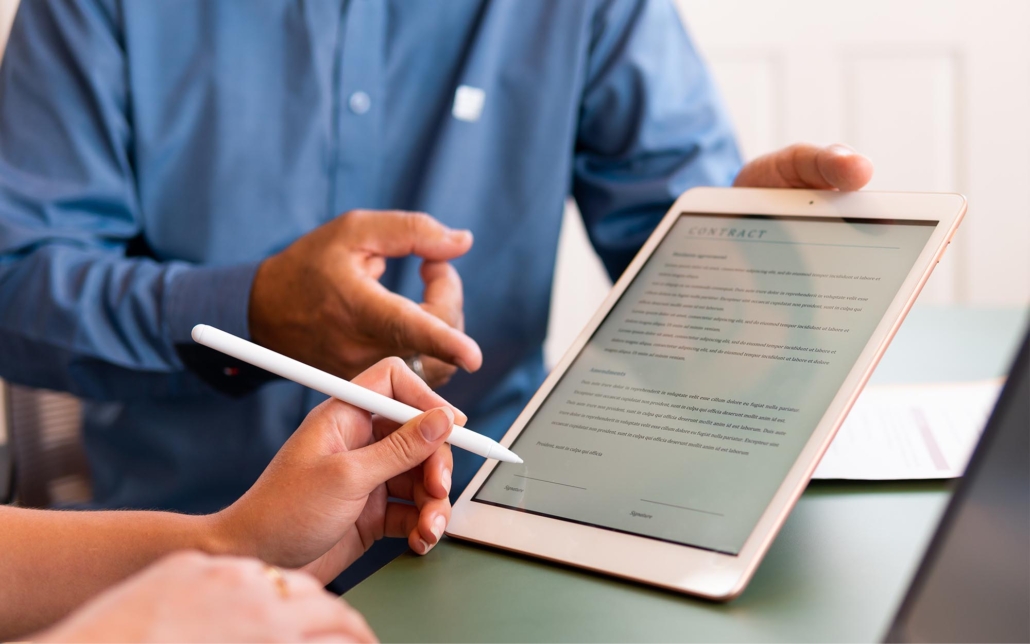 Two individuals review a contract on a tablet, with one person pointing to specific sections while the other holds a stylus, ready to sign. Their focused interaction highlights the importance of careful document review and digital signing in modern business practices.