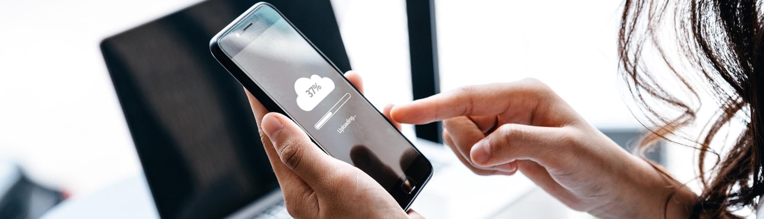 A woman backs up her mobile device to the cloud, carefully navigating the settings on her phone. Her focused expression and the display indicating data transfer highlight the importance of securing her information and ensuring it's safely stored online.