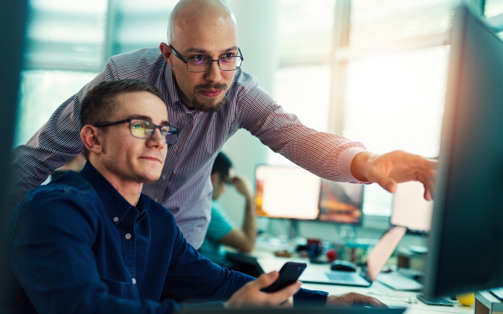 A manager assists an employee in troubleshooting a problem on a desktop computer. The manager provides guidance and support, leaning in to offer advice while the employee attentively follows along. Their collaborative effort reflects a commitment to resolving the issue efficiently and fostering a supportive work environment.