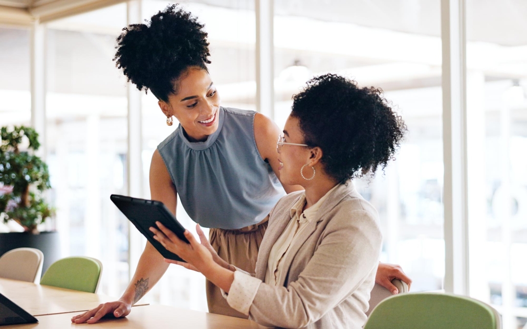 Two office workers enjoy a lively conversation while looking at an iPad, sharing smiles and engaging in animated discussion. Their positive interaction and the collaborative use of technology reflect a productive and friendly office environment.