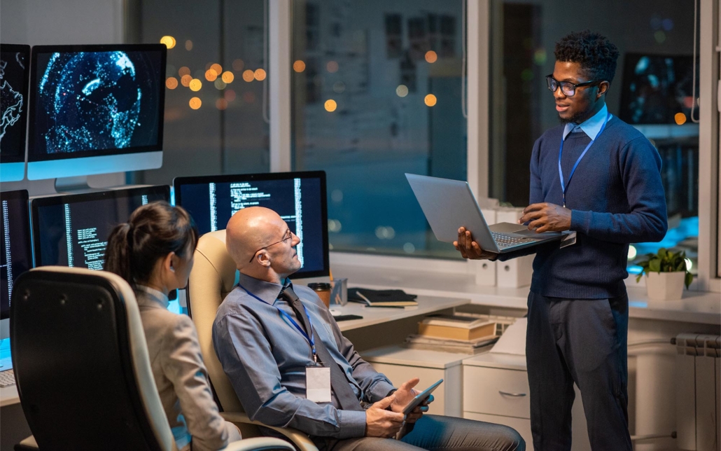 Three business professionals engage in cyber training, attentively following a presentation or instructor. They sit around with laptops and tablets, demonstrating a collaborative approach to learning about cybersecurity practices and strategies. Their focused expressions and active participation underscore their commitment to enhancing their knowledge and skills in safeguarding digital assets.