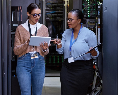 Two professional women work together in a server room, facing forward as they manage and monitor network equipment. Their focused expressions and coordinated efforts reflect a commitment to maintaining and optimizing server performance in a high-tech environment.