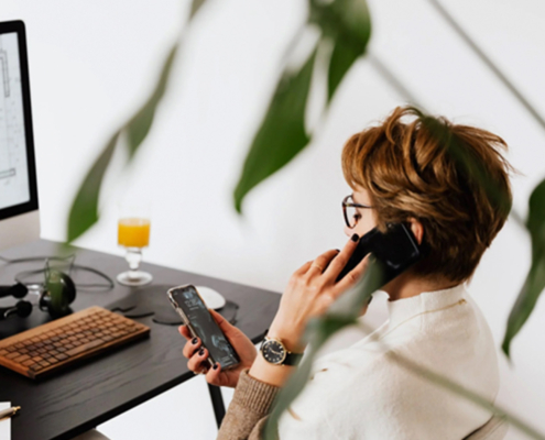 A businessman multitasks with efficiency, holding a phone to his ear while engaging in a conversation on another phone placed in front of his laptop. His focused demeanor and organized workspace suggest a busy, productive environment.