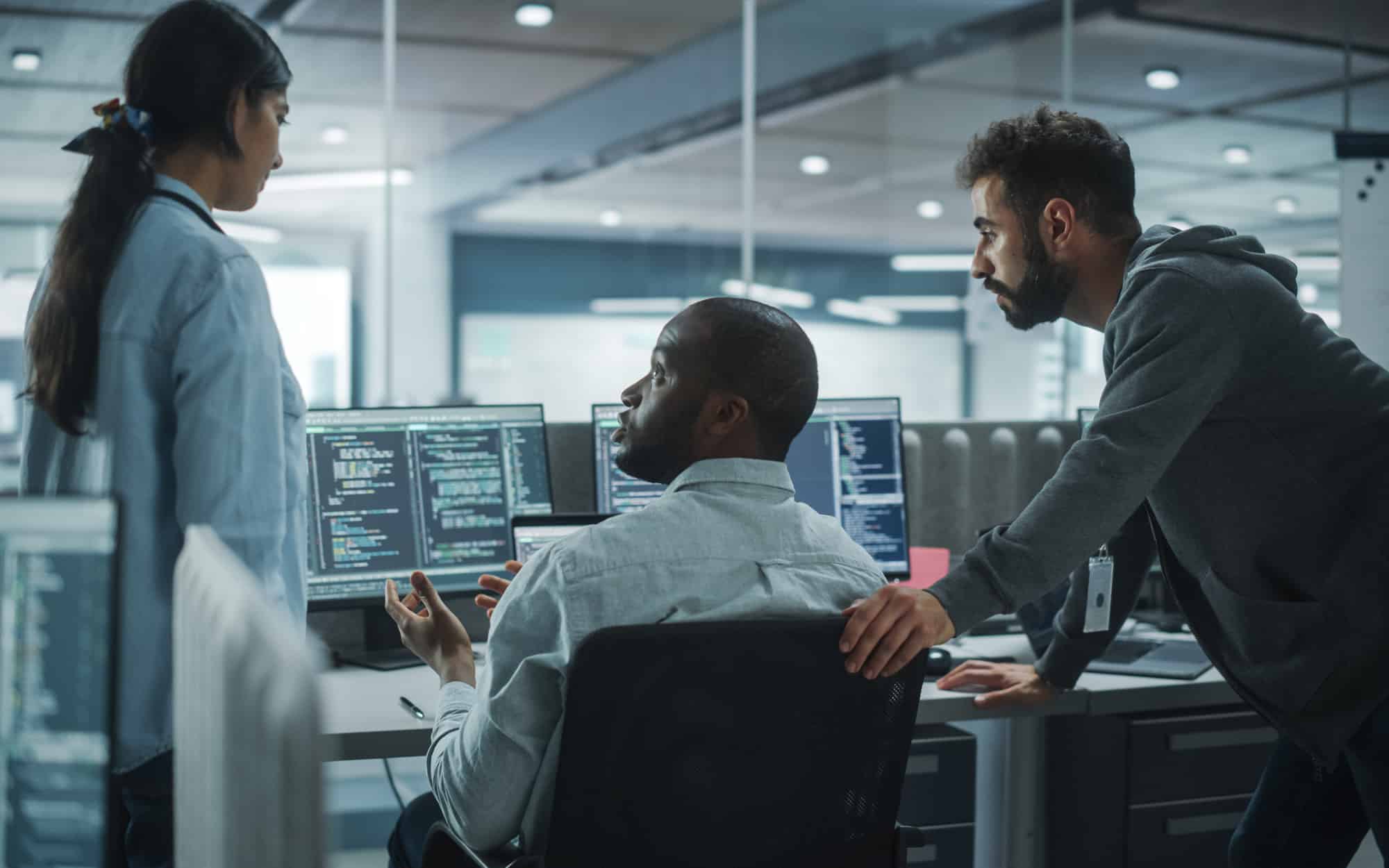 A diverse group of IT professionals collaborating in a modern office environment, surrounded by computer screens displaying code and data. The team is engaged in a discussion, focused on solving complex technical issues, showcasing teamwork and innovation in the tech industry.