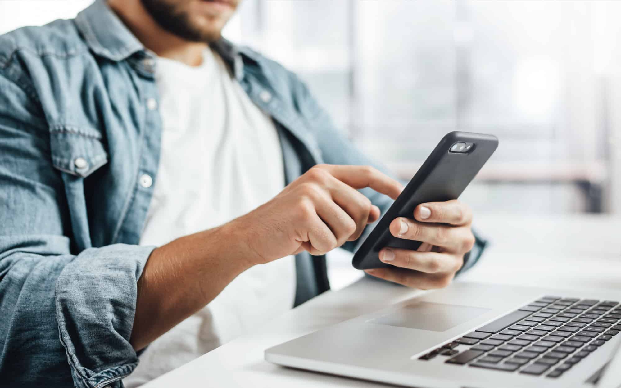 A focused professional sits at his sleek, modern desk, engaged in a multitasking feat. With one hand, he expertly navigates his smartphone, scrolling through emails and updates. His other hand is busy typing on a keyboard, managing ongoing tasks with precision.