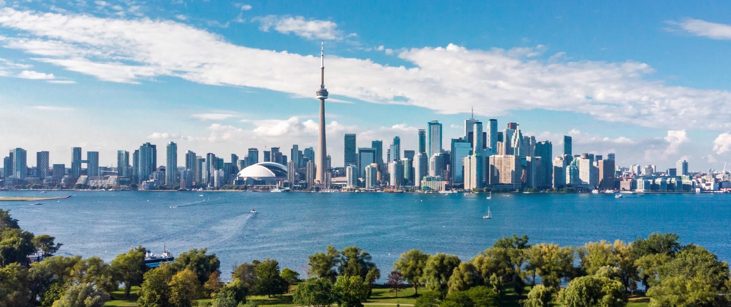 A panoramic view of Toronto's skyline, featuring the iconic CN Tower and Rogers Centre, with the city's skyscrapers lining the waterfront. The scene is framed by lush green parks in the foreground and the tranquil waters of Lake Ontario under a bright blue sky with scattered clouds.