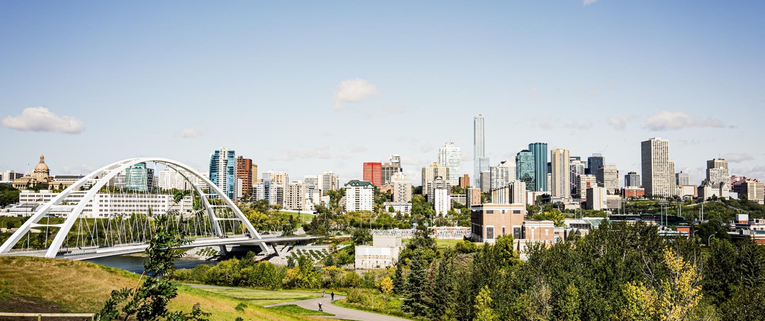 A scenic view of Edmonton's city skyline featuring modern high-rise buildings, lush greenery, and a prominent white arch bridge under a clear blue sky.