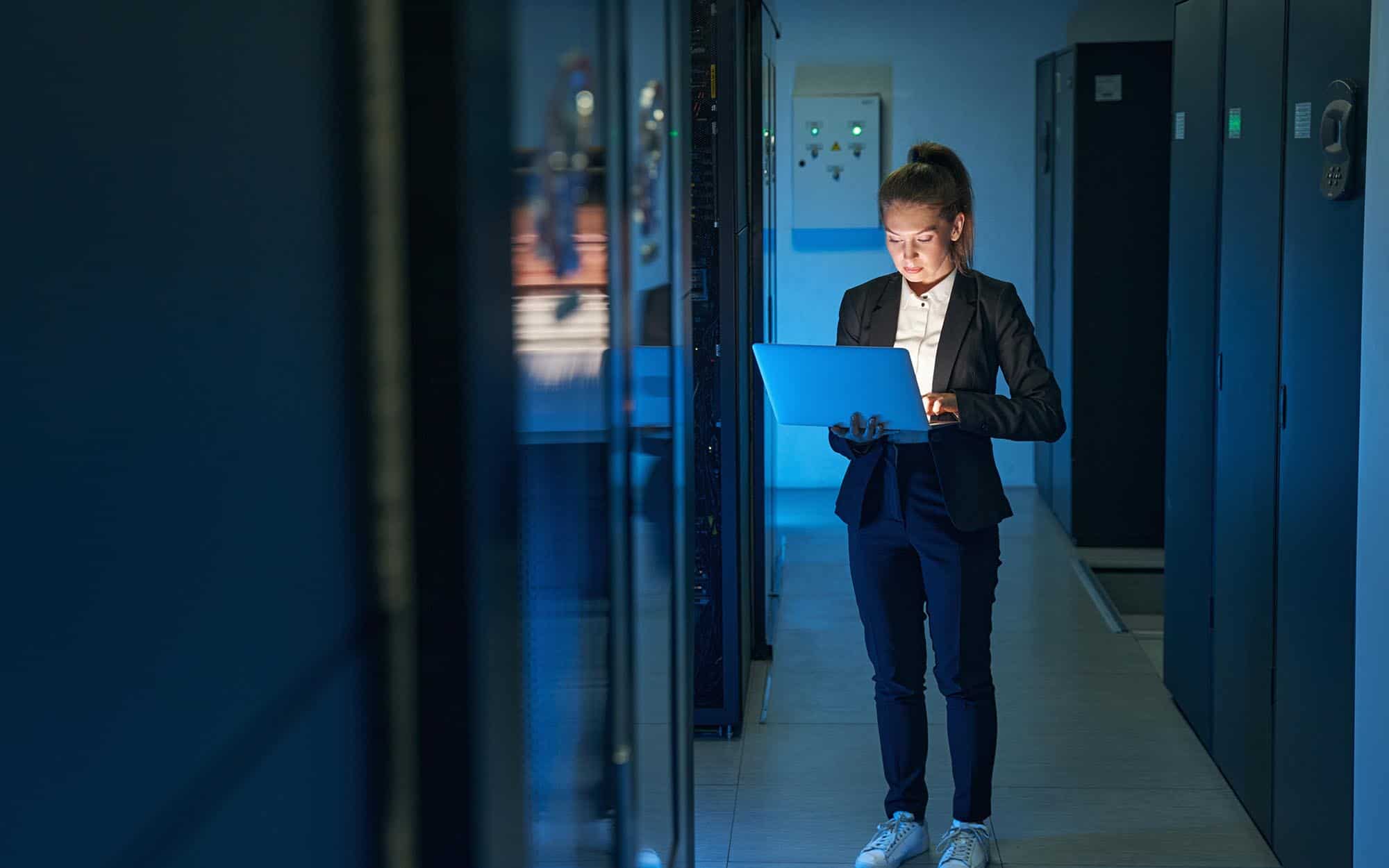 A woman in a server room working on a laptop, surrounded by server racks and networking equipment, highlighting IT infrastructure and network management.