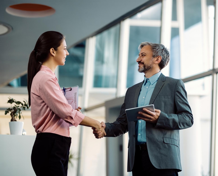 A businesswoman and a businessman shaking hands in a modern office setting, both holding documents, symbolizing a successful professional agreement or partnership.