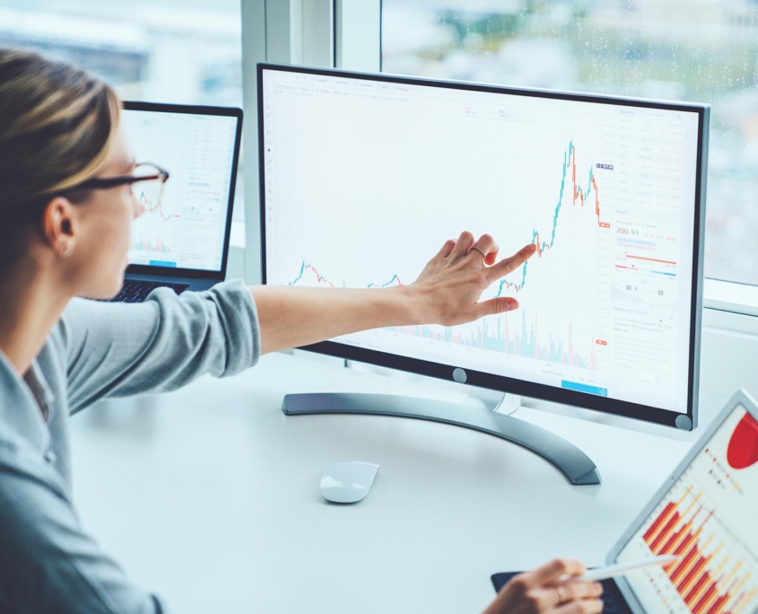 A woman pointing at a graph on a computer monitor, with another person analyzing charts on a tablet and a laptop in the background, illustrating data analysis and financial review.